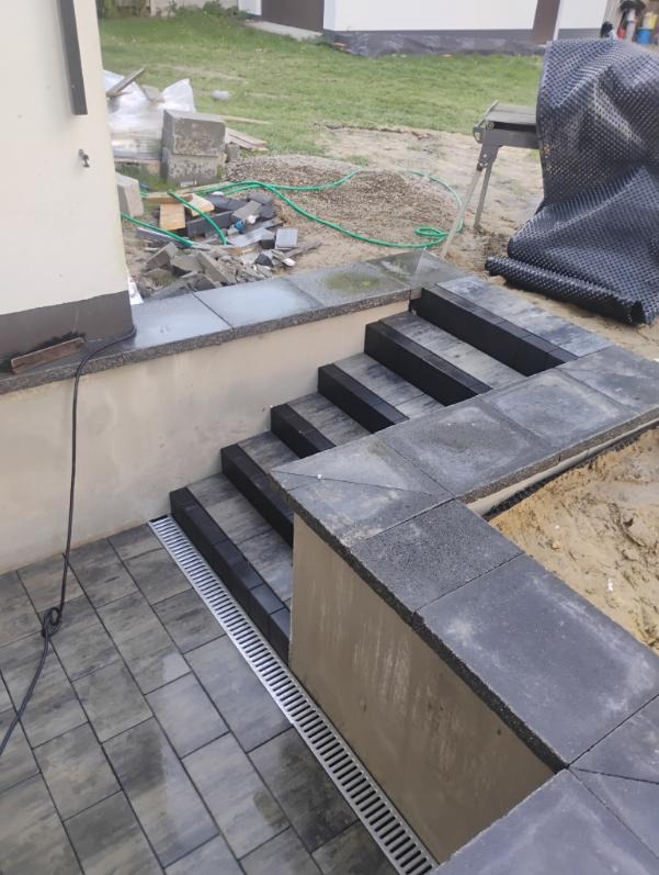 Nowe schody z kostki brukowej w odcieniach szarości, z widocznym odpływem liniowym i elementami przygotowanymi do dalszych prac wykończeniowych na zewnątrz budynku.