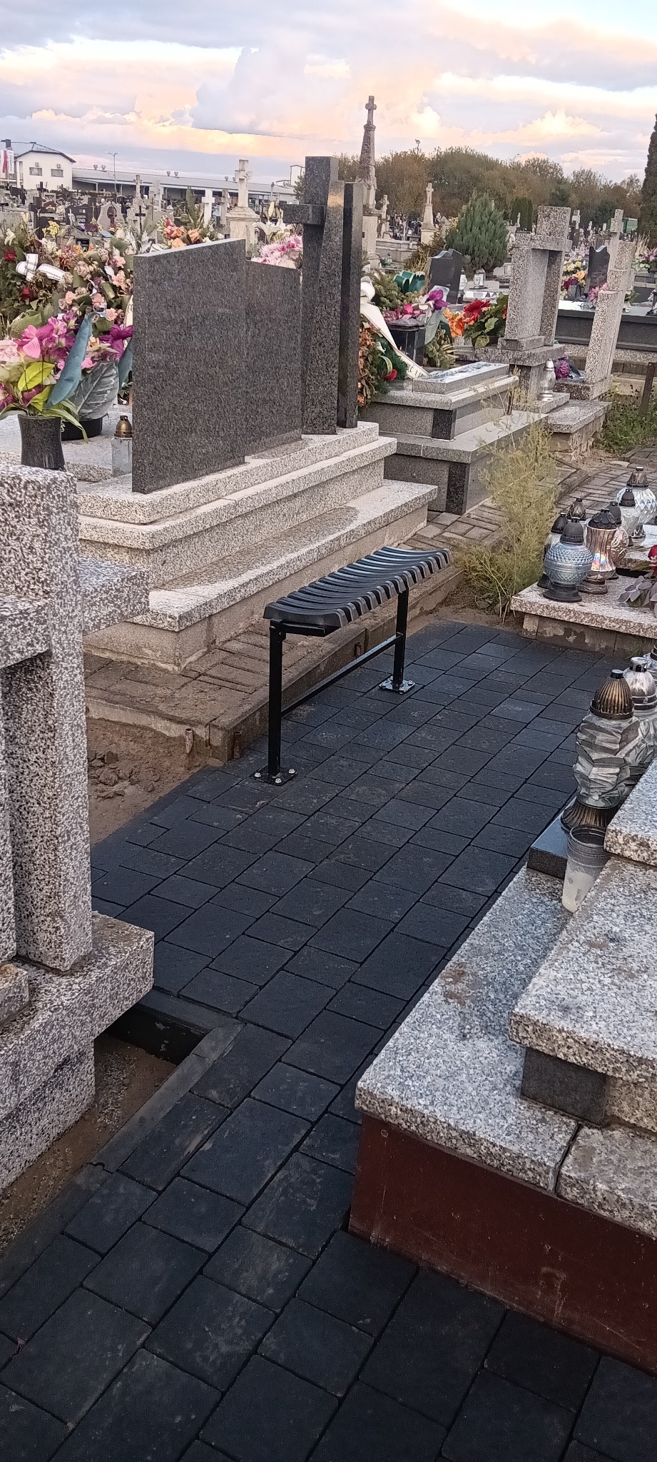 Ciemna kostka brukowa ułożona na cmentarzu między nagrobkami, z czarną ławką. Widok z góry, oświetlenie naturalne, nagrobki z kwiatami w tle.