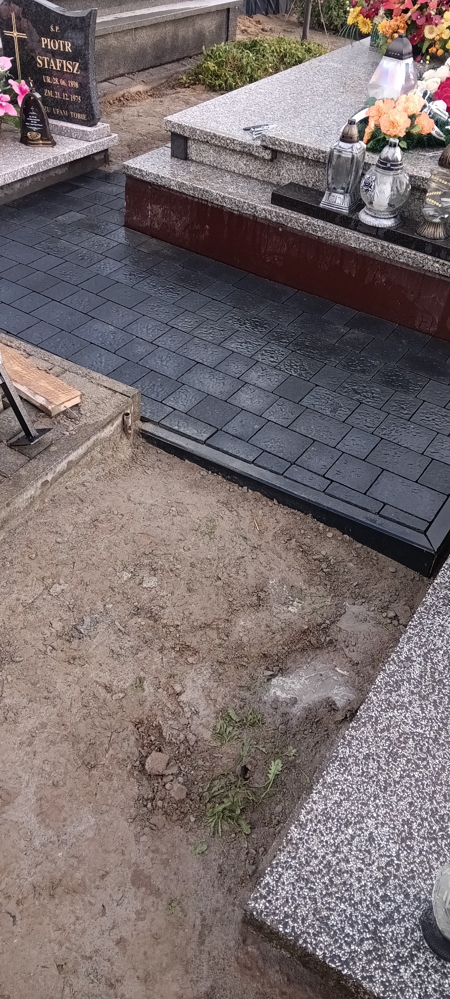 Częściowo ułożona, ciemna kostka brukowa wokół nagrobka z szarego granitu. Fragment wykopu z ziemią. Widoczne kwiaty i znicze na nagrobku.