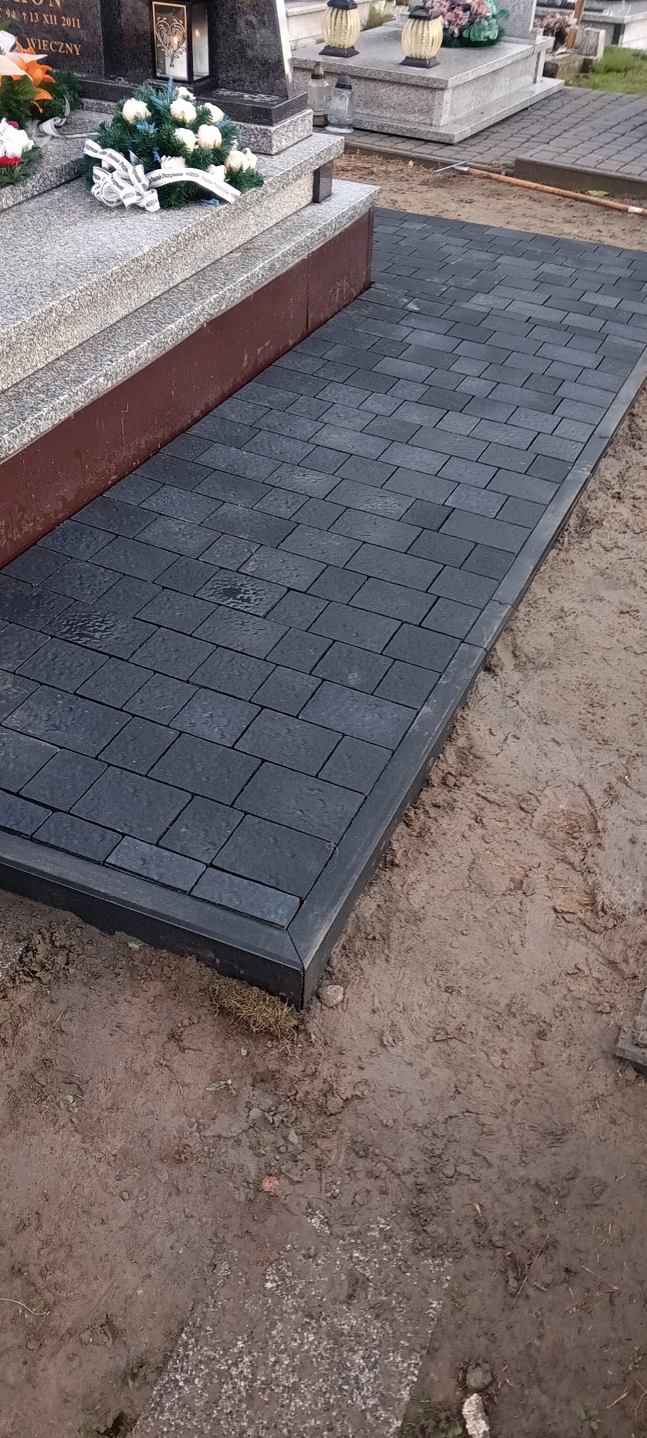 Ułożona, grafitowa kostka brukowa przy nagrobku, z widocznym obramowaniem. Świeżo ułożona nawierzchnia na cmentarzu, ziemia wokół.