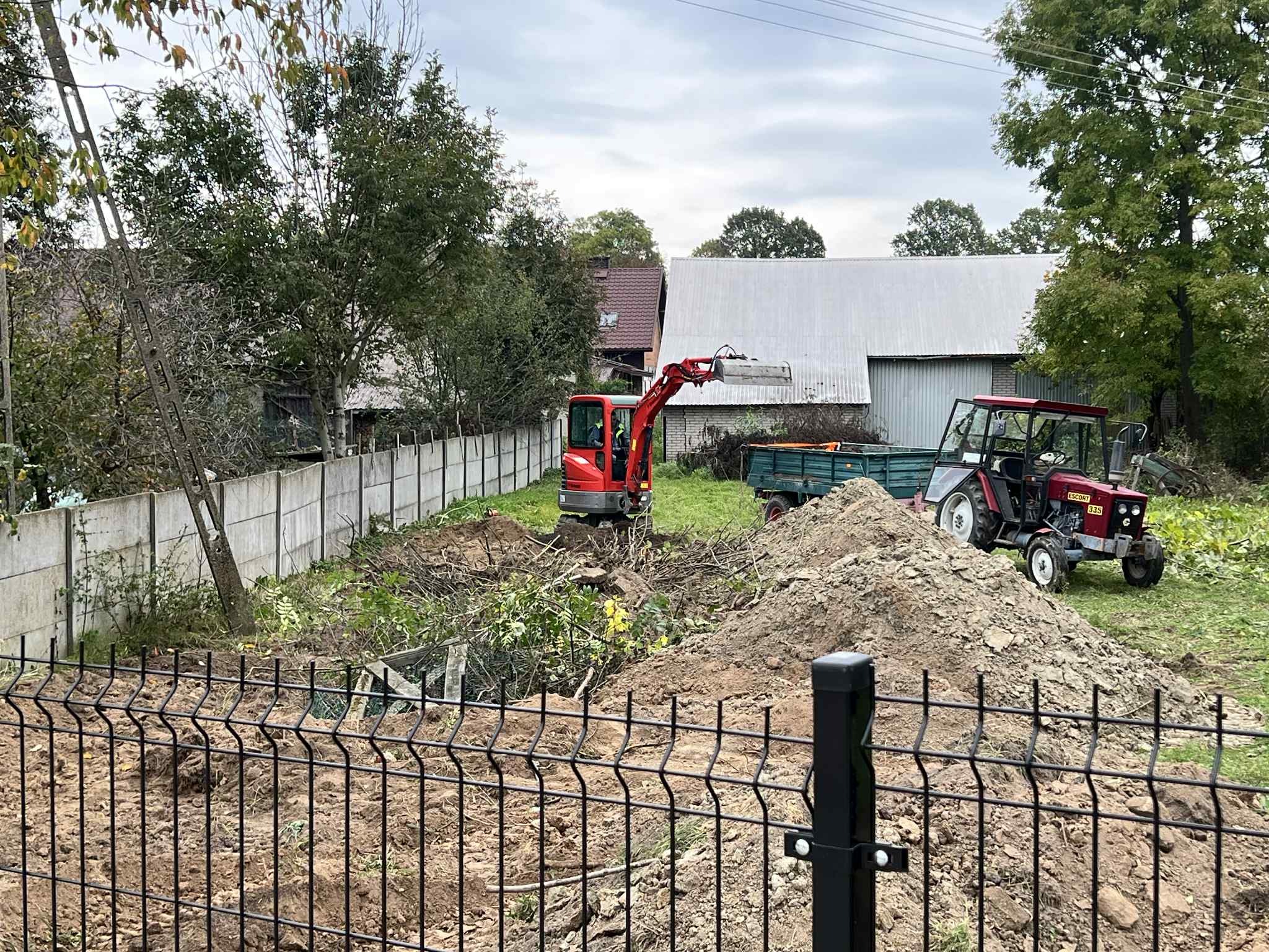 Czerwony mini-koparka i traktor z przyczepą na działce w trakcie niwelacji, widoczne ogrodzenie panelowe i betonowe. Prace ziemne w toku, przygotowanie terenu pod budowę.