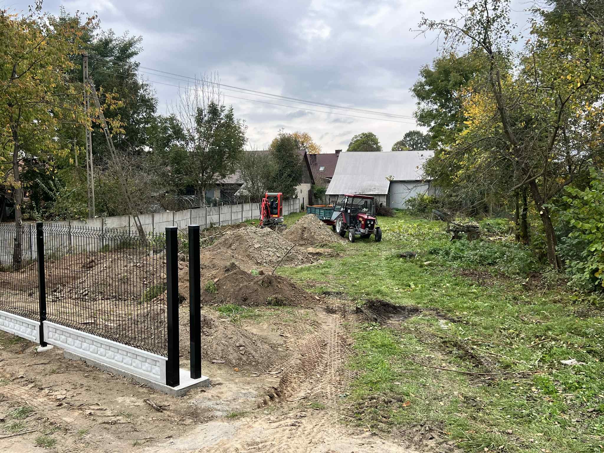 Montaż ogrodzenia panelowego z podmurówką na nierównym terenie. W tle widoczny traktor i mała koparka. Pochmurne niebo.