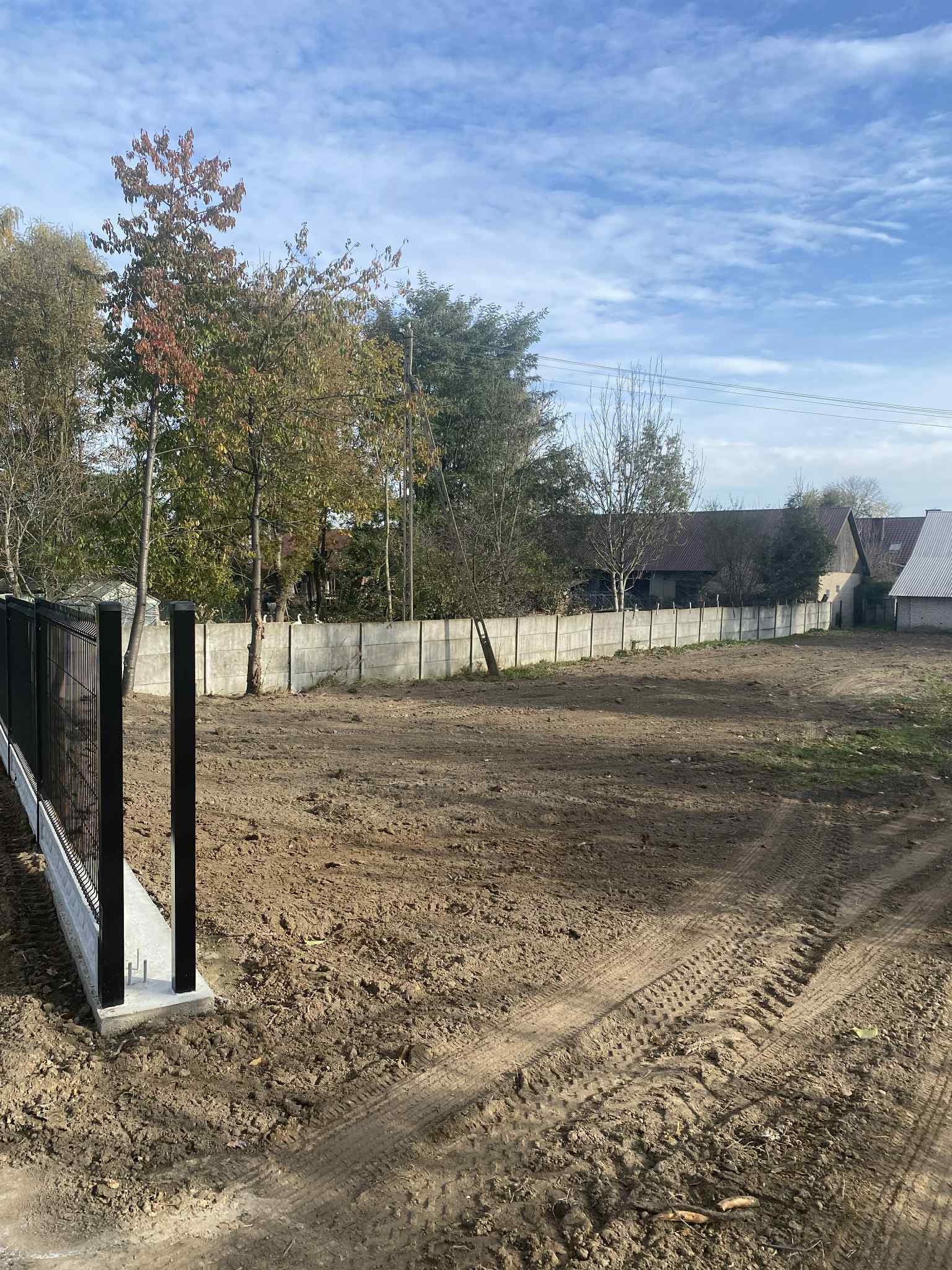 Nowoczesne ogrodzenie betonowe z elementami metalowymi, świeżo zamontowane na nierównym terenie. Widoczne ślady opon, drzewa w tle i błękitne niebo.