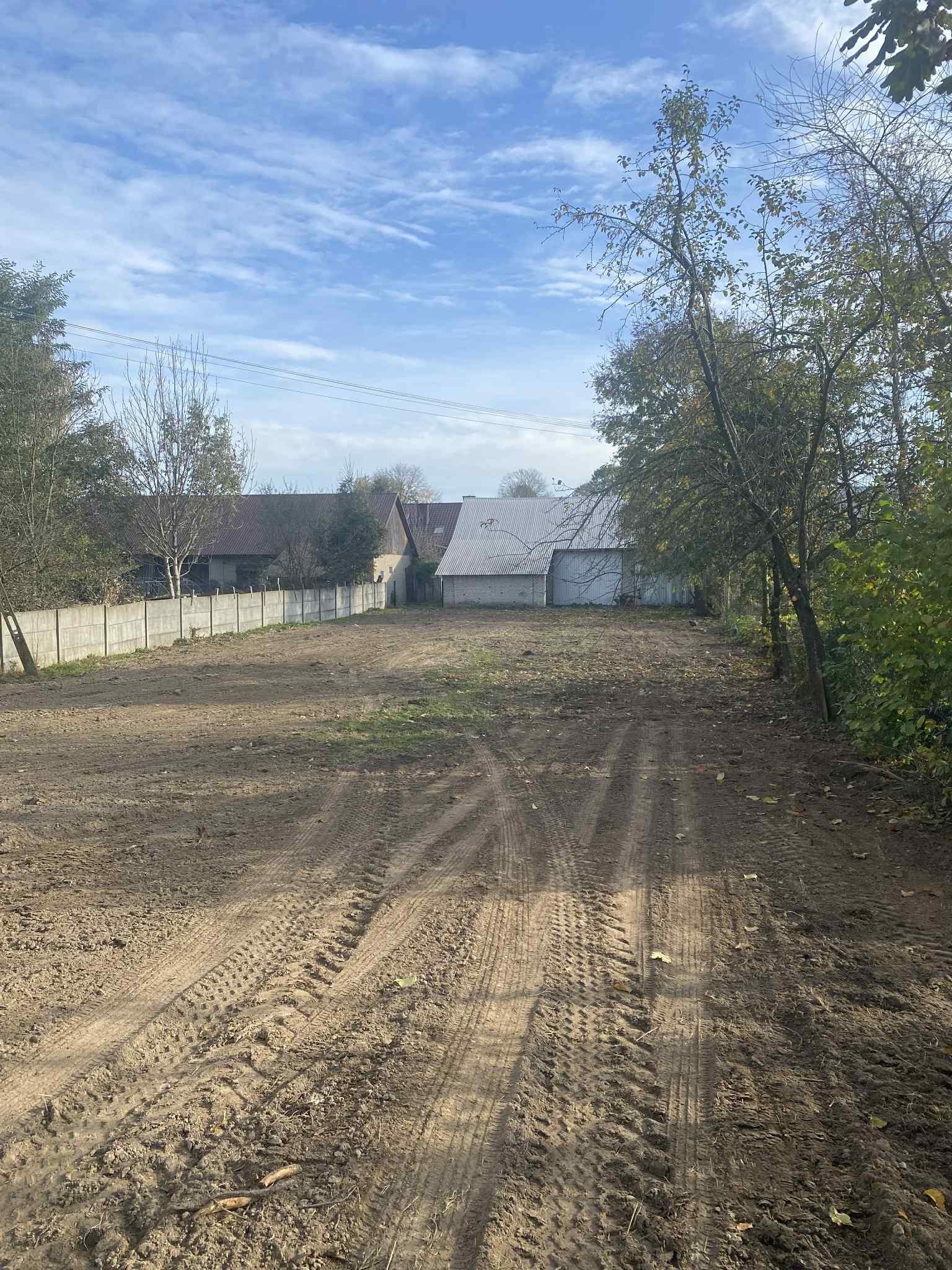 Przygotowany teren pod budowę z widocznymi śladami opon, otoczony drzewami i betonowym ogrodzeniem. W tle budynki gospodarcze pod błękitnym niebem.