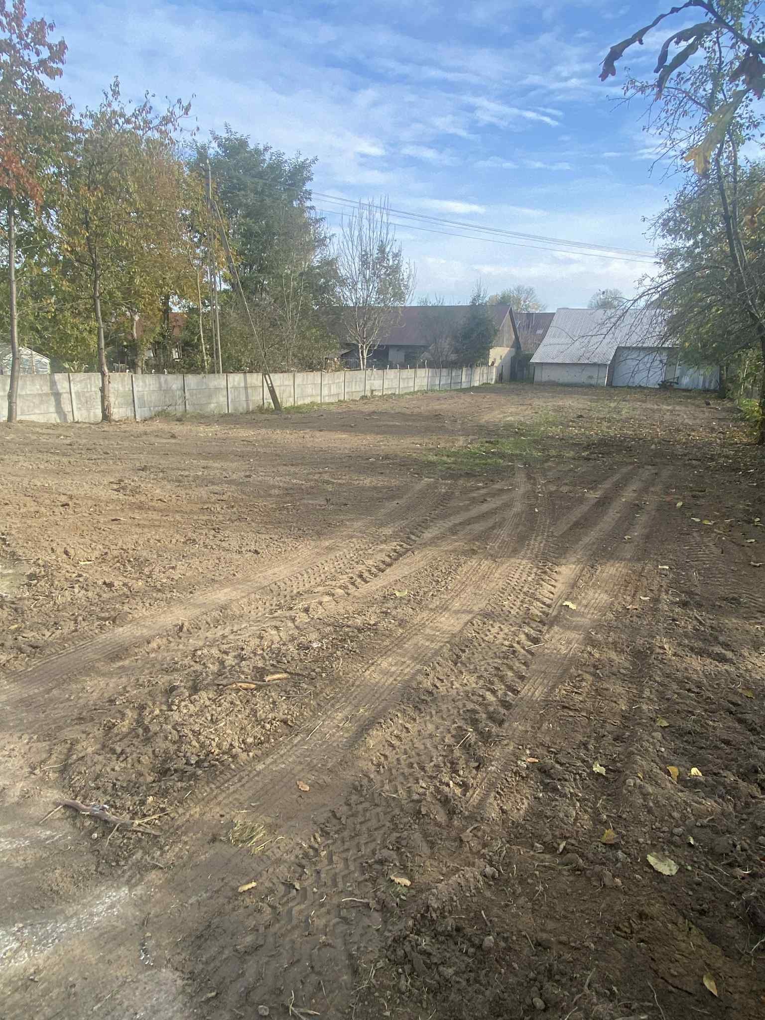 Przygotowany teren pod budowę z widocznymi śladami opon, otoczony betonowym ogrodzeniem. W tle budynki gospodarcze i drzewa pod błękitnym niebem.