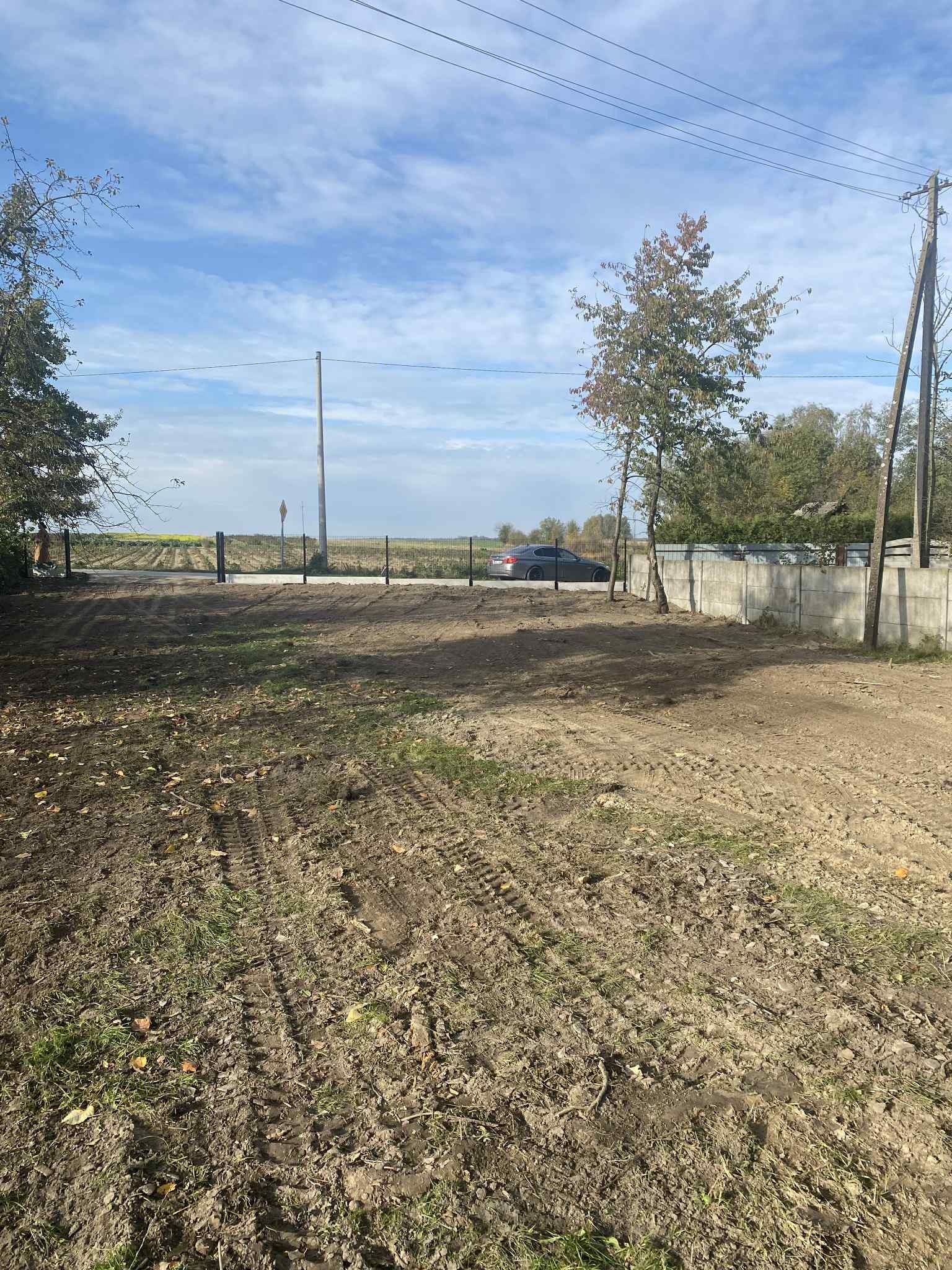 Działka po niwelacji z widocznymi śladami opon, częściowo ogrodzona siatką i betonowym płotem. W tle pole i błękitne niebo z chmurami.