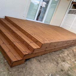 Vasyl Yatsko - Nowo wybudowany, brązowy taras z drewna, z czterostopniowymi schodami, przylegający do jasnej ściany budynku z białymi oknami i drewnianą bramką z kratką w tle.