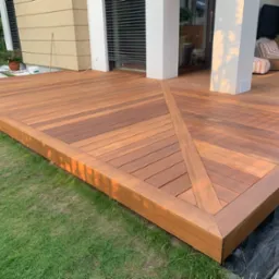 Elegancki taras z drewna egzotycznego, z geometrycznym wzorem ułożenia desek. Widok na dom z dużymi oknami i meblami ogrodowymi. Starannie wykonane wykończenie.