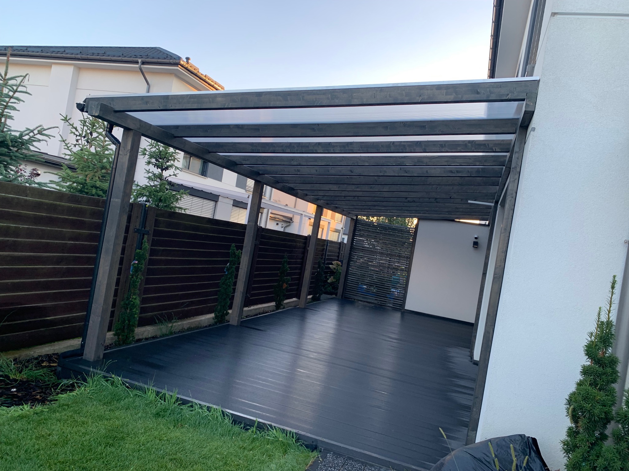 Zadaszona przestrzeń z ciemną podłogą i drewnianą konstrukcją, widok na ogród i sąsiedni budynek, nowoczesny design.