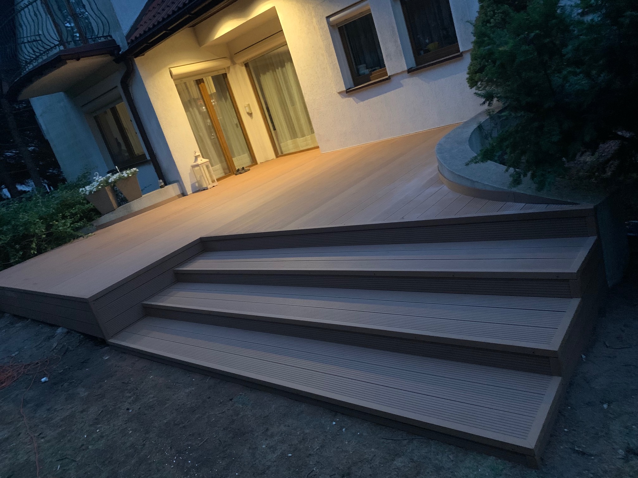 Nowoczesny taras kompozytowy z czterostopniowymi schodami prowadzącymi do domu, oświetlony ciepłym światłem z wnętrza, otoczony zielenią.