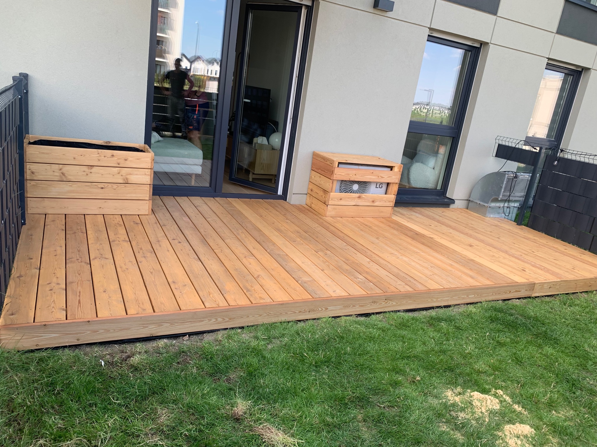 Nowoczesny taras z jasnego drewna z drewnianymi donicami i obudową klimatyzatora, widok na trawnik i budynek mieszkalny.