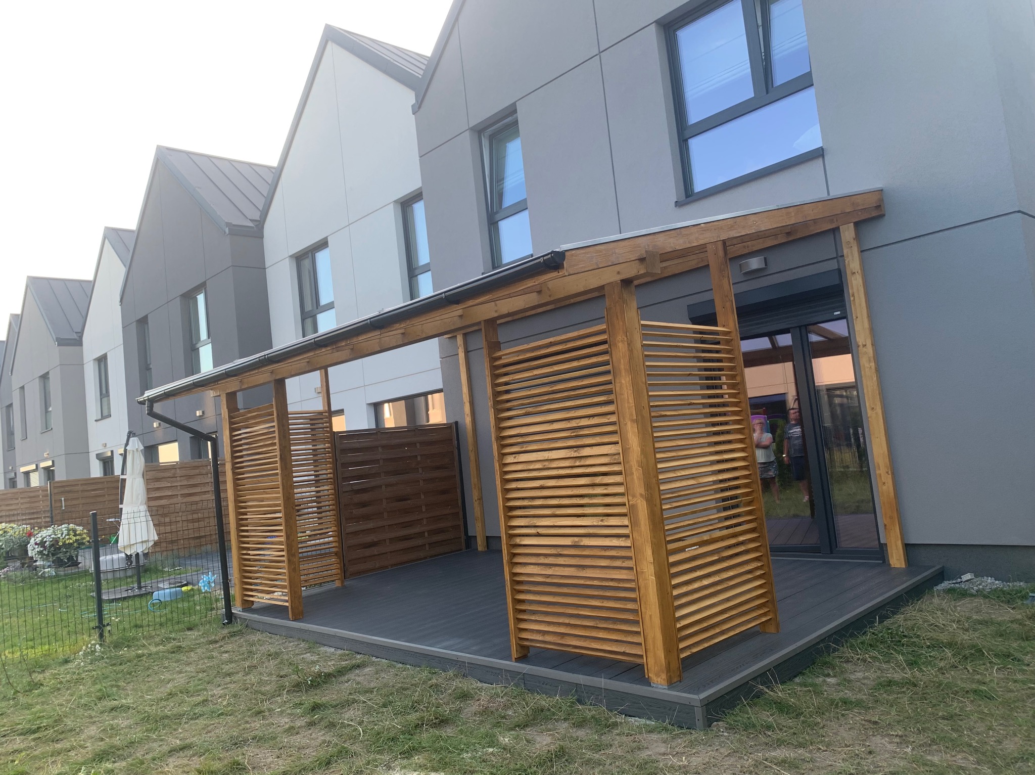 Nowoczesna pergola drewniana z lamelami na tarasie kompozytowym, przylegająca do szarego domu o minimalistycznej architekturze, widok z trawnika.