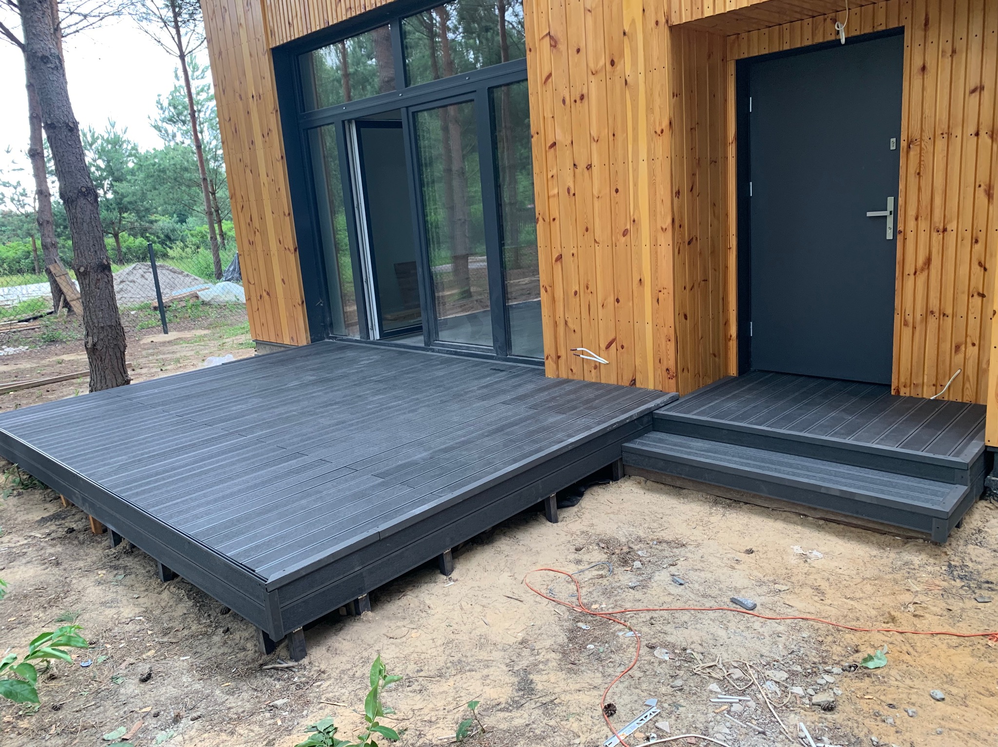 Nowoczesny dom z elewacją z jasnego drewna i tarasem z desek kompozytowych w kolorze grafitowym, widoczne schody wejściowe i przesuwne drzwi tarasowe, otoczenie w stanie surowym.