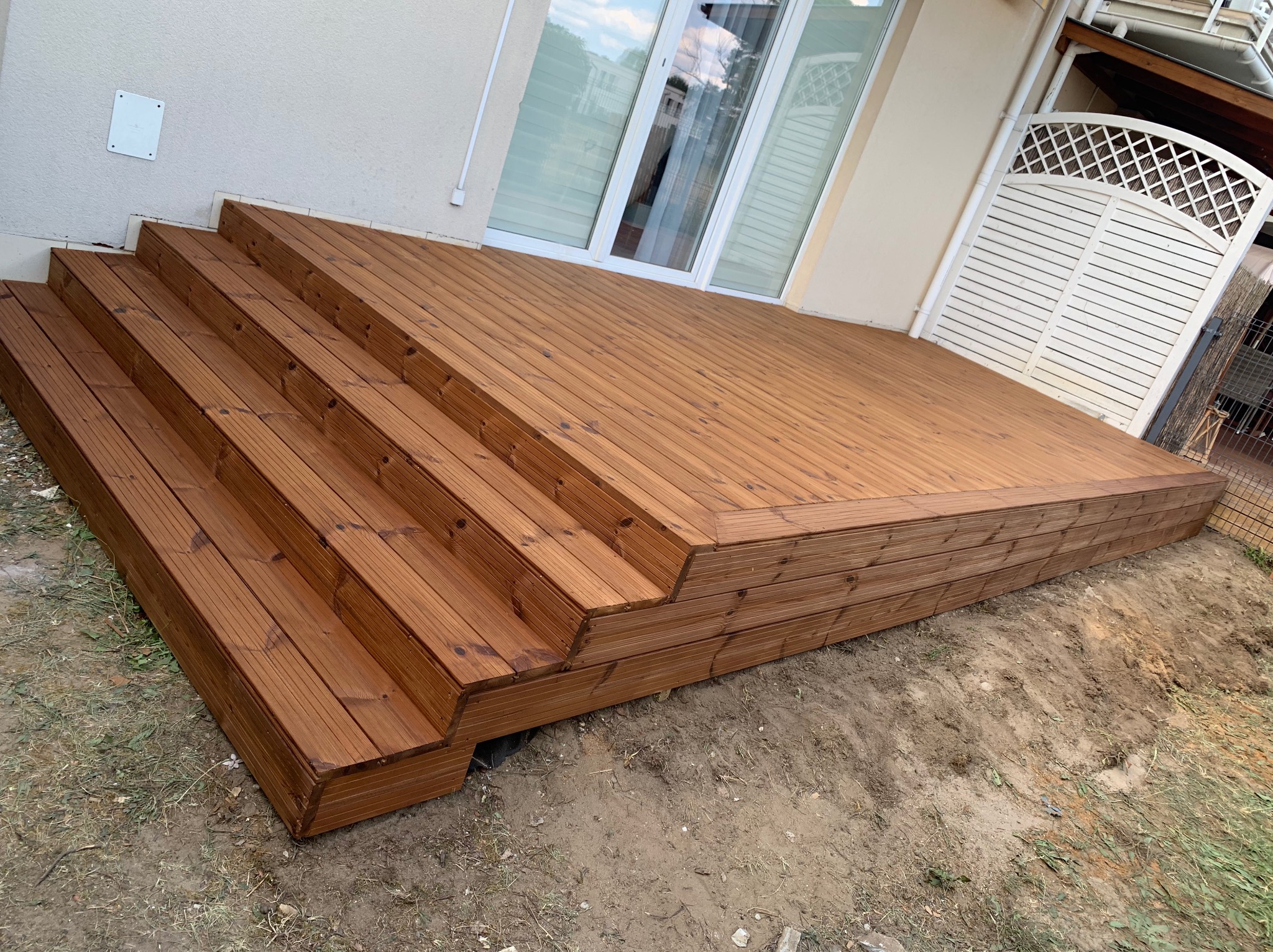 Nowo wybudowany, brązowy taras z drewna, z czterostopniowymi schodami, przylegający do jasnej ściany budynku z białymi oknami i drewnianą bramką z kratką w tle.