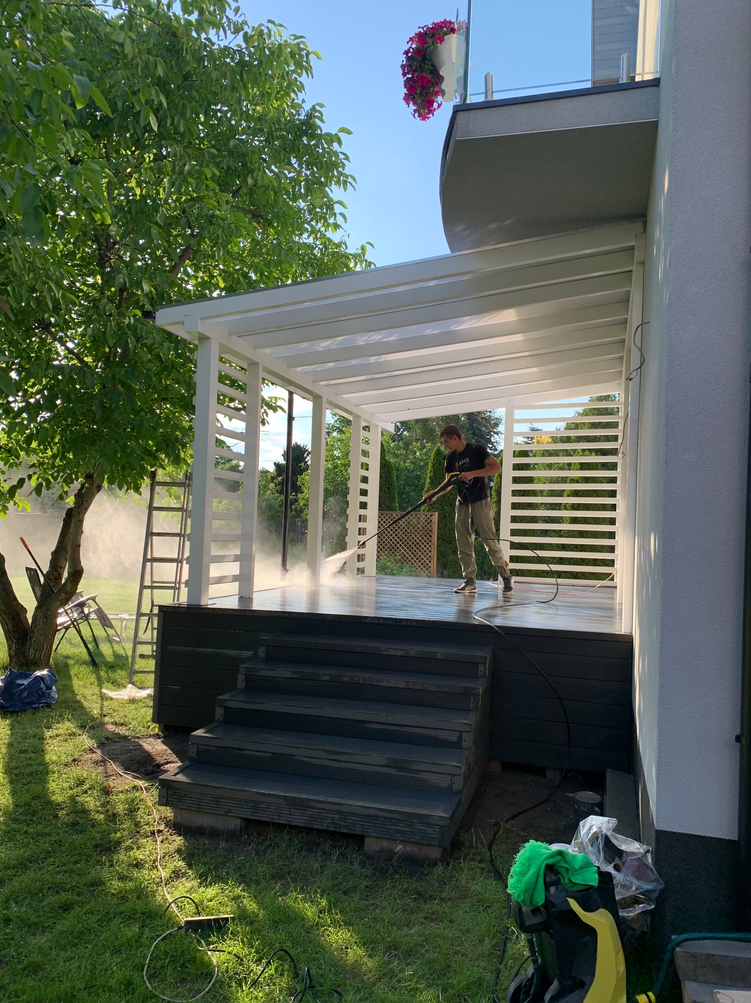 Czyszczenie pod wysokim ciśnieniem powierzchni pod białą, nowoczesną pergolą z drewnianymi schodami wejściowymi; widoczna drabina oparta o drzewo i akcesoria do czyszczenia na trawniku.