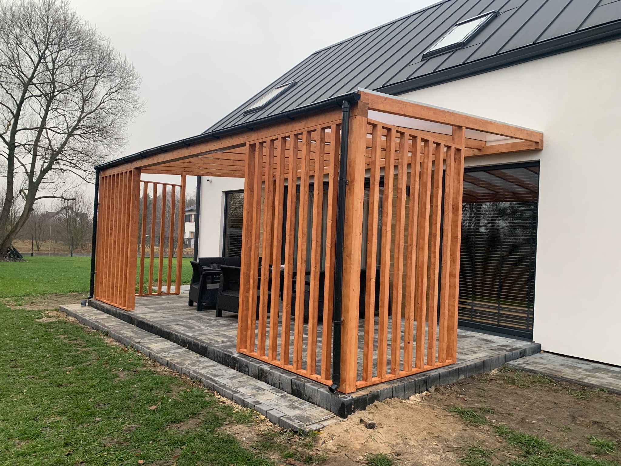 Nowoczesna pergola z jasnego drewna z pionowymi listwami, przylegająca do białego domu z czarnym dachem, z widocznym fragmentem tarasu wyłożonego kostką brukową i czarnymi meblami ogrodowymi wewnątrz.