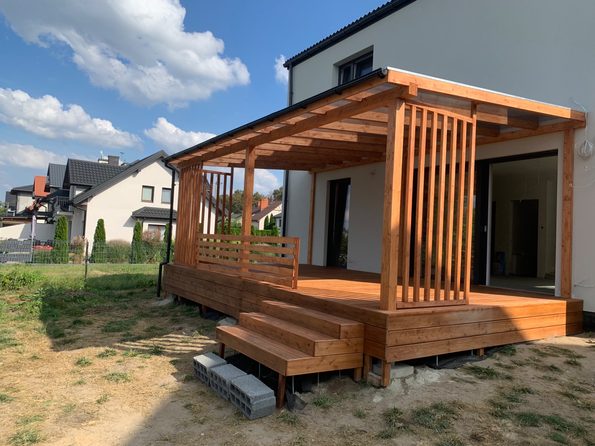 Drewniany taras z pergolą przy białym domu w słoneczny dzień. Widoczne schody i elementy konstrukcyjne. Tło: domy i niebieskie niebo z chmurami.
