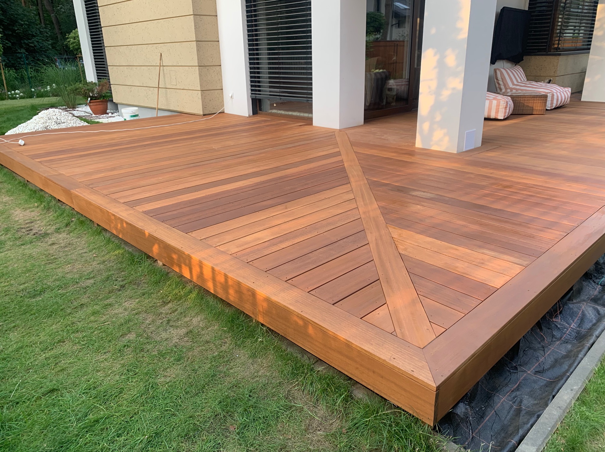 Elegancki taras z drewna egzotycznego, z geometrycznym wzorem ułożenia desek. Widok na dom z dużymi oknami i meblami ogrodowymi. Starannie wykonane wykończenie.