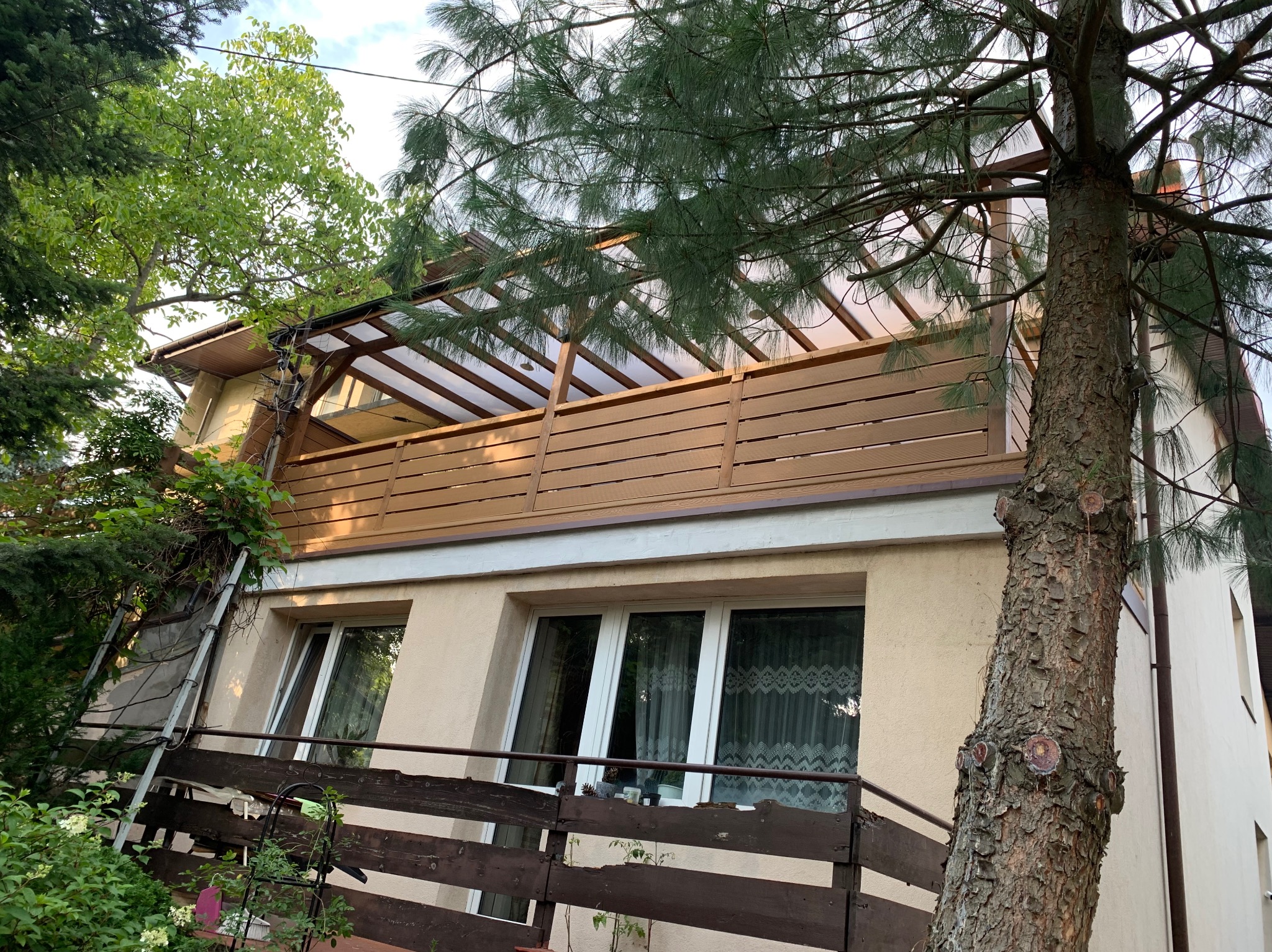 Zadaszony balkon z drewnianą balustradą na piętrze domu, widoczny z dołu, otoczony zielenią drzew. Na balkonie widoczna konstrukcja z przezroczystym dachem.