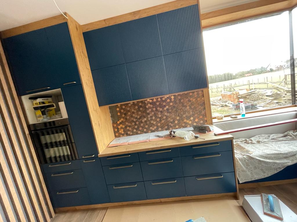 Niebieska zabudowa kuchenna z fornirowanymi frontami i mozaiką nad blatem, widok na okno i plac budowy w tle, w trakcie montażu.