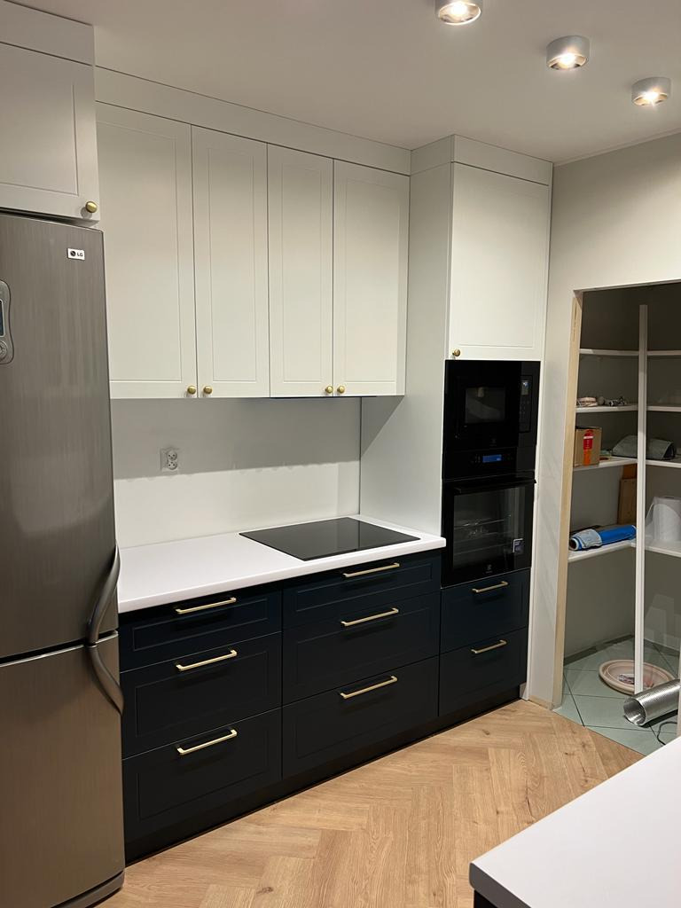 Nowoczesna kuchnia: białe górne szafki z okrągłymi, złotymi uchwytami, ciemnogranatowe dolne szafki z długimi, złotymi uchwytami, biały blat, płyta indukcyjna, zabudowany piekarnik, podłoga w jodełkę.