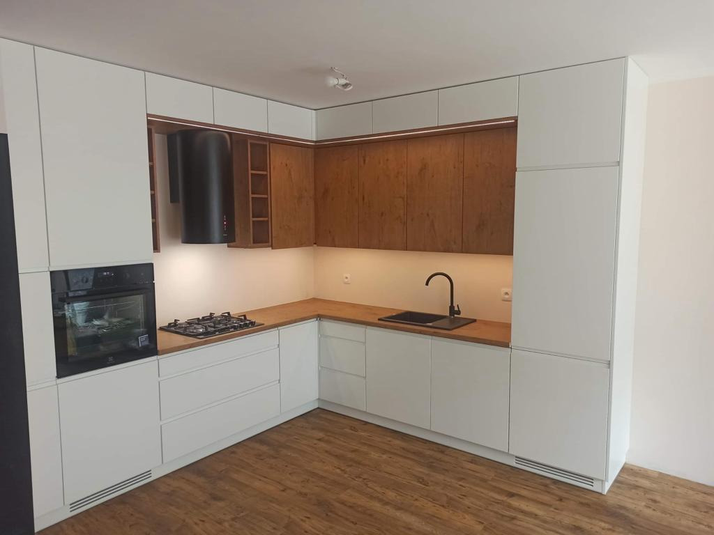 Nowoczesna kuchnia w kształcie litery L z białymi frontami i drewnianymi górnymi szafkami, czarny okap nad płytą gazową, drewniany blat i podłoga.