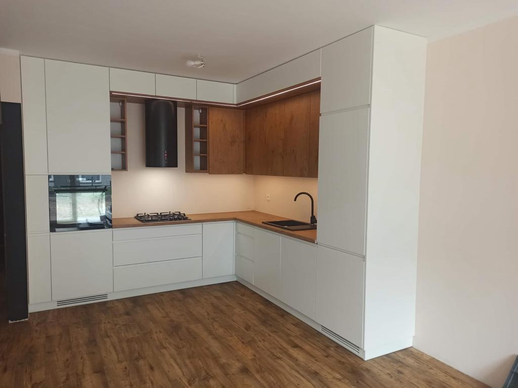 Nowoczesna kuchnia w kształcie litery L z białymi frontami i drewnianymi akcentami, oświetleniem LED pod szafkami wiszącymi i drewnianą podłogą.