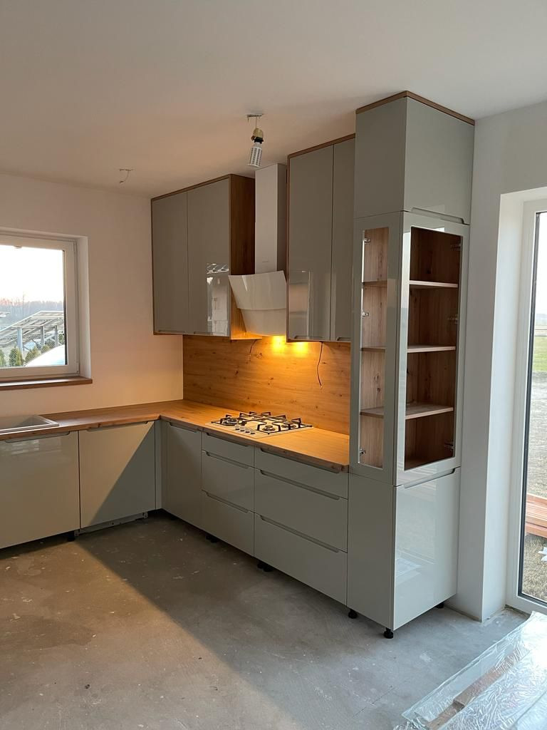 Jasnoszara kuchnia narożna z drewnianym blatem i okładziną ścienną, widok na płytę gazową i przeszkloną szafkę z półkami, surowy beton na podłodze.