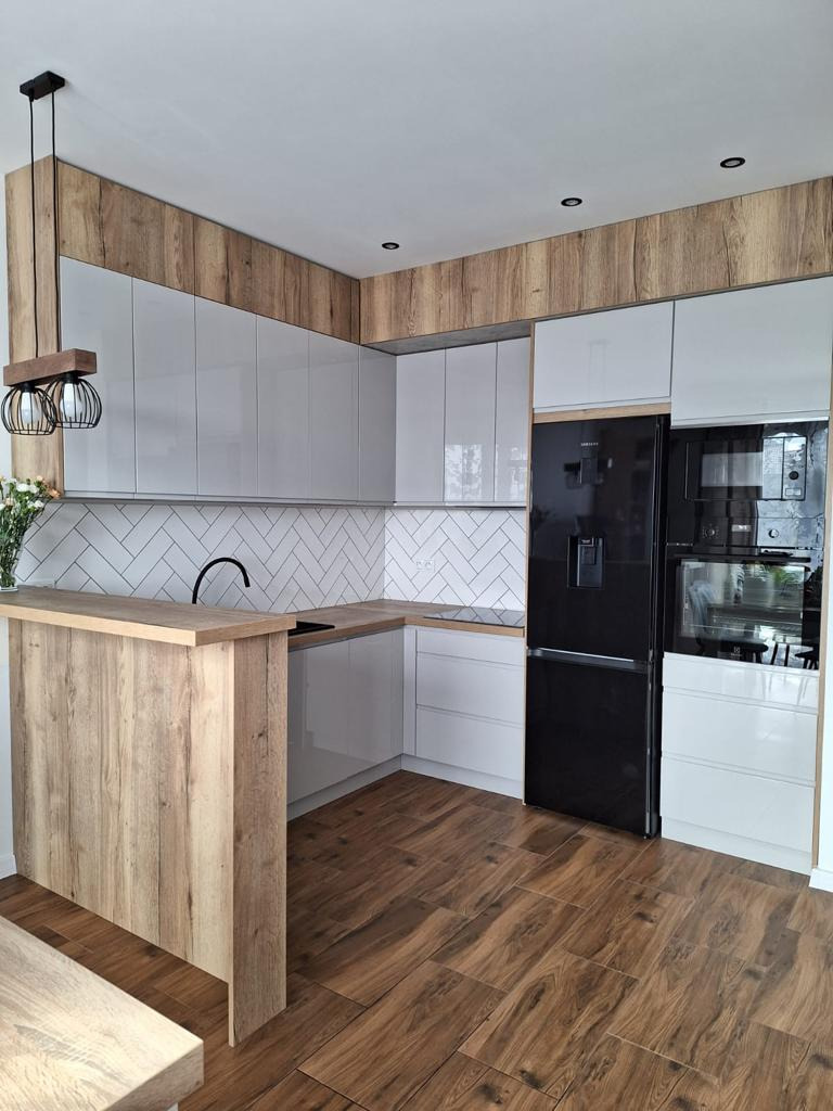 Nowoczesna kuchnia z białymi, lakierowanymi szafkami, drewnopodobną zabudową górną i wyspą, czarną lodówką i płytkami w jodełkę na ścianie.
