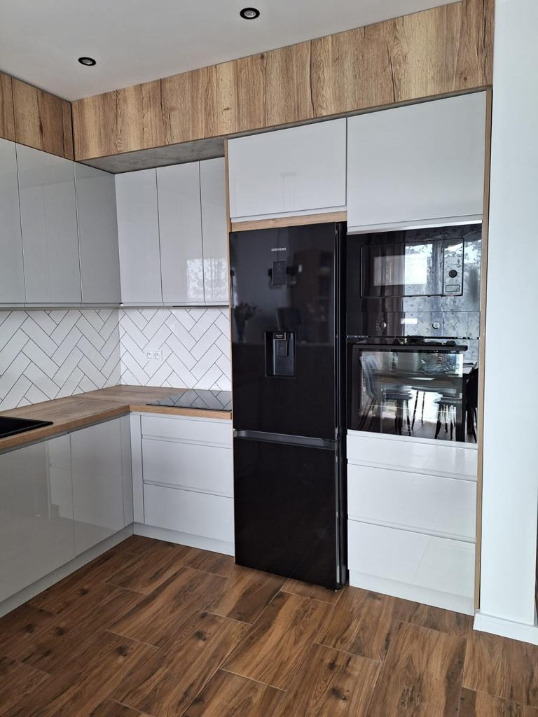 Nowoczesna kuchnia z białymi, błyszczącymi frontami szafek, drewnianymi akcentami na górnych szafkach i blacie, oraz czarną lodówką w zabudowie i piekarnikiem. Na ścianie białe płytki ułożone...