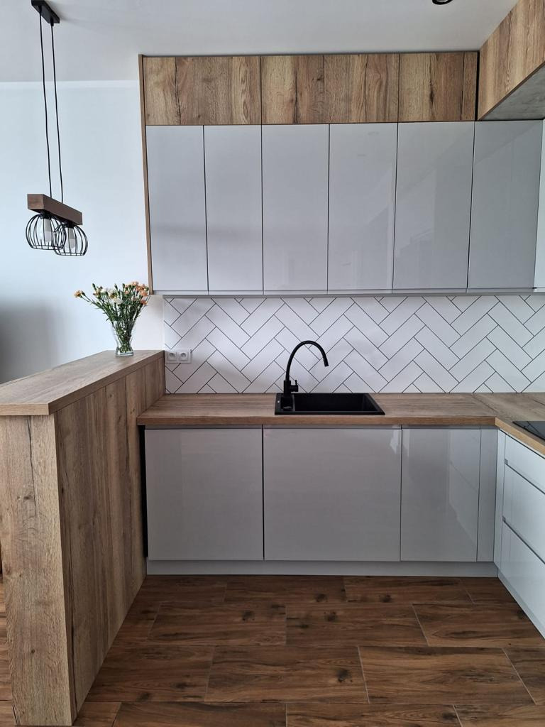 Jasnoszara kuchnia z drewnianymi akcentami, czarnym zlewem i baterią, oraz białymi kafelkami ułożonymi w jodełkę, oświetlona designerską lampą wiszącą.
