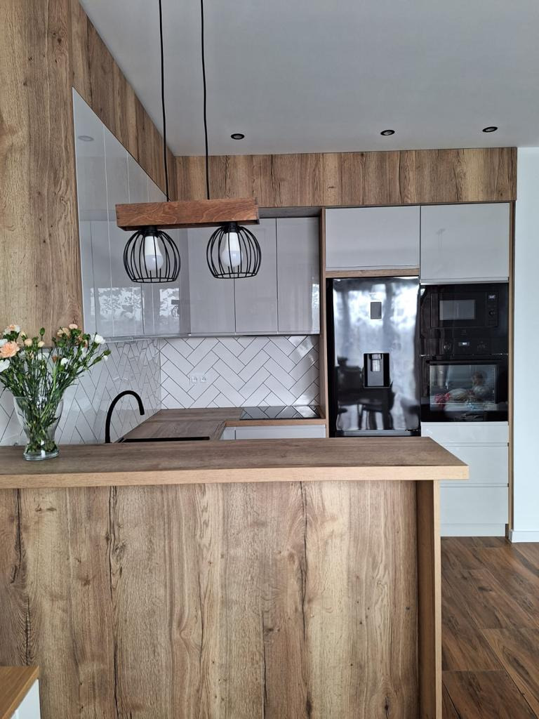 Nowoczesna kuchnia z drewnianym blatem wyspowym, szare szafki, białe płytki w jodełkę na ścianie, oświetlenie wiszące z drewnianą belką i czarnymi kloszami.