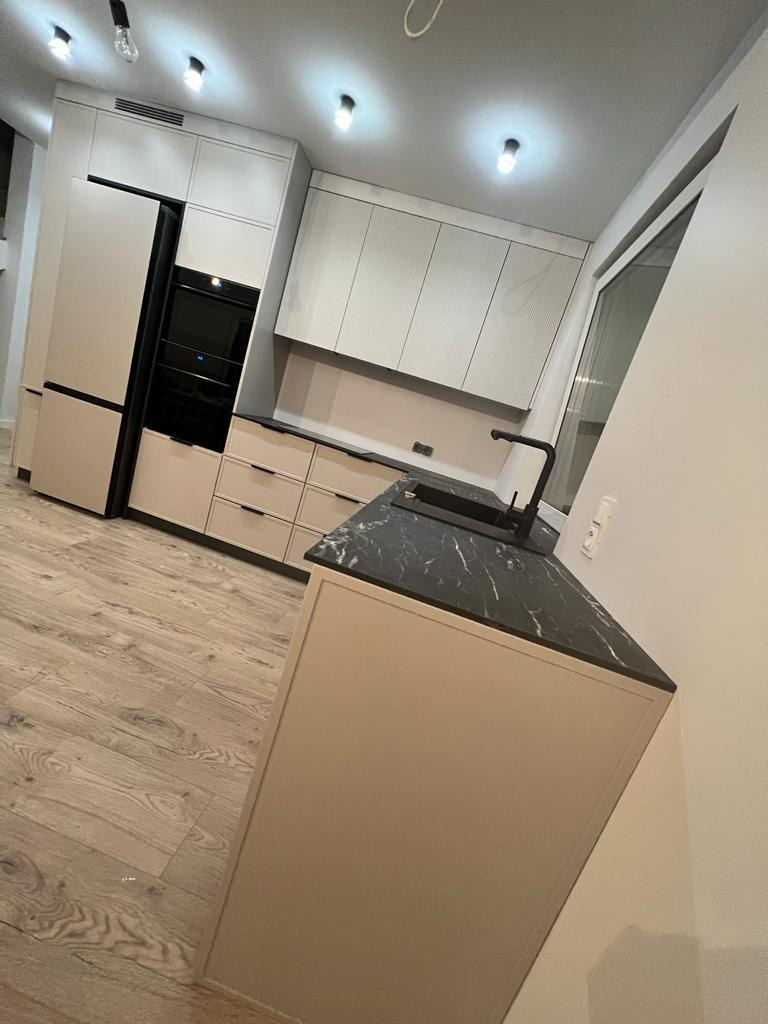 Nowoczesna kuchnia z beżowymi frontami szafek, czarnym blatem z marmurowym wzorem i wbudowanym sprzętem AGD, oświetlona punktowymi lampami sufitowymi.