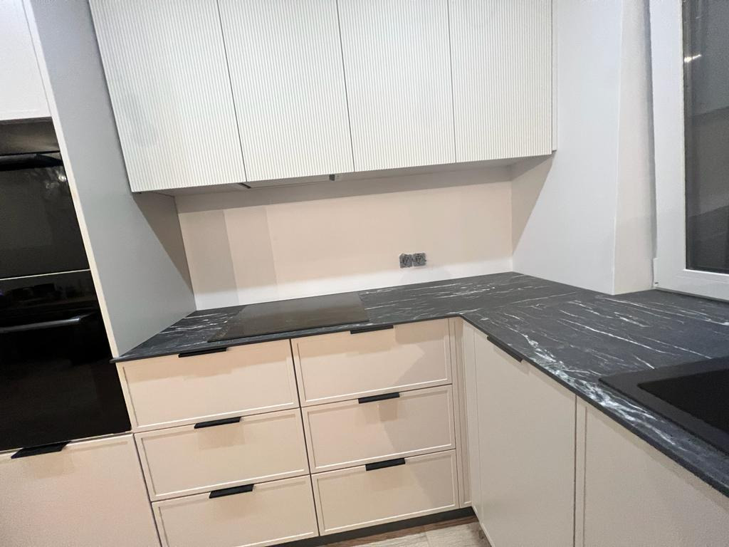 Nowoczesna kuchnia z beżowymi szafkami z szufladami, czarnym blatem imitującym marmur i białymi szafkami górnymi z ryflowanym frontem, widok na narożnik kuchenny z płytą grzewczą i zlewem.