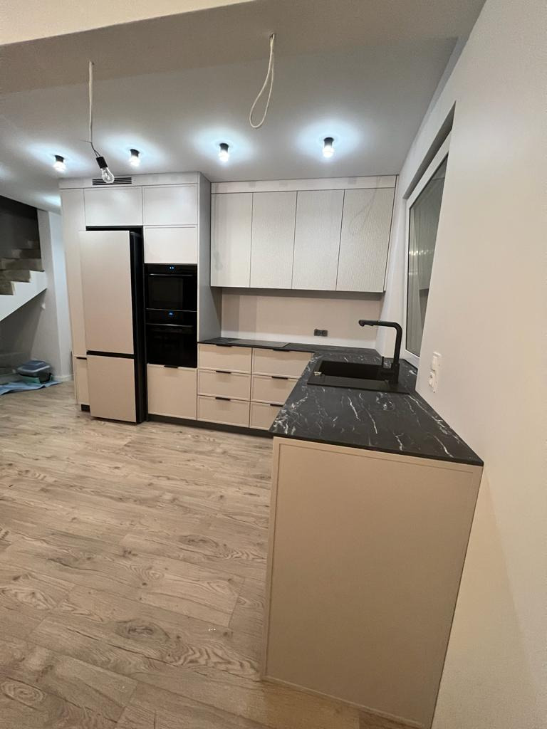 Nowoczesna kuchnia z beżowymi frontami, czarnym blatem z marmurowym wzorem i sprzętem AGD w zabudowie. Niedokończone oświetlenie sufitowe z widocznymi kablami.