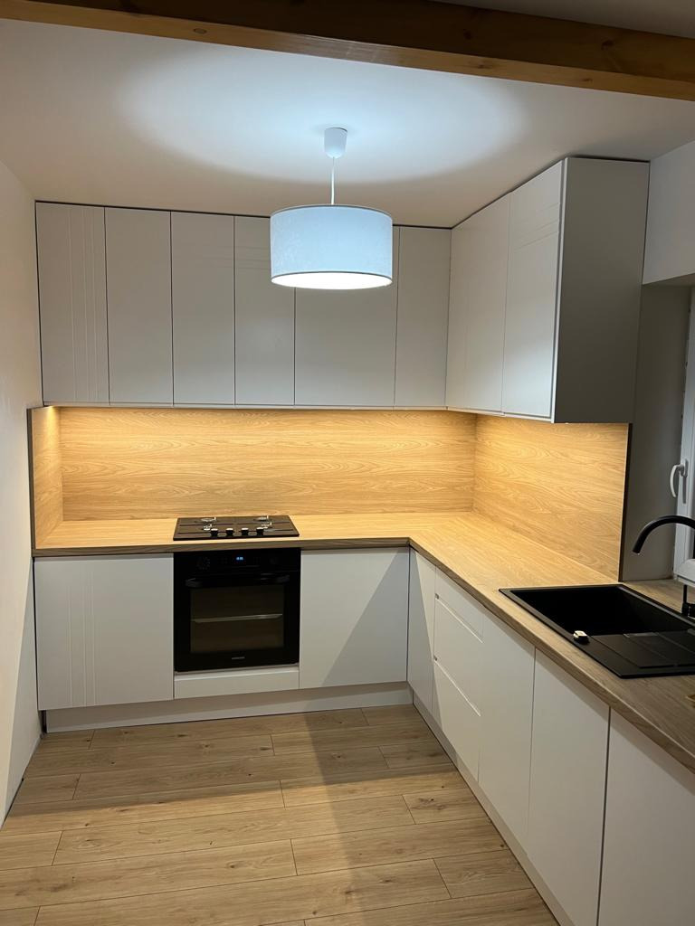 Nowoczesna kuchnia w kształcie litery L z białymi frontami, drewnianym blatem i backsplashiem w kolorze jasnego drewna, oświetlona wiszącą lampą z abażurem i punktowym światłem pod szafkami.