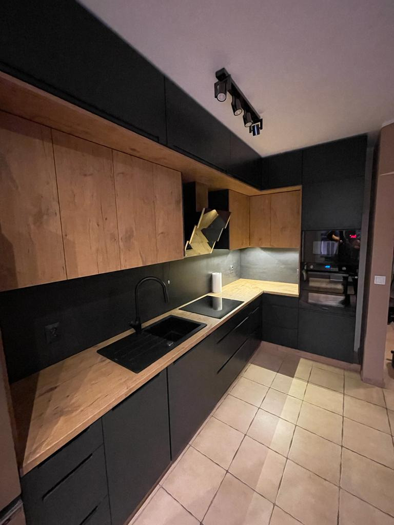 Nowoczesna kuchnia z czarnymi szafkami dolnymi i drewnianymi górnymi, czarny zlew i bateria, oświetlenie punktowe na suficie, płytki podłogowe w odcieniach beżu.