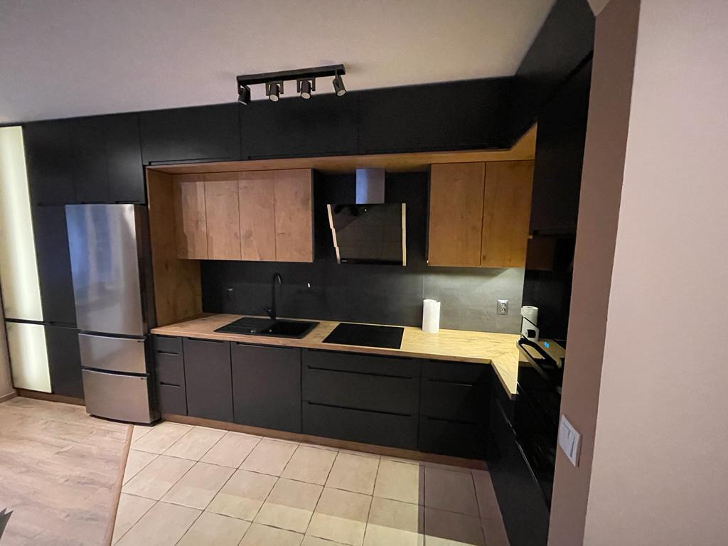 Nowoczesna kuchnia z czarnymi frontami szafek dolnych i drewnianymi górnymi, oświetlenie punktowe, srebrna lodówka, płytki podłogowe w odcieniach beżu.