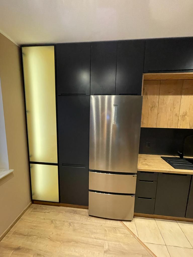 Nowoczesna kuchnia z czarnymi szafkami, złotymi frontami i lodówką ze stali nierdzewnej, widok na blat z drewnianym dekorem i czarnym zlewem.