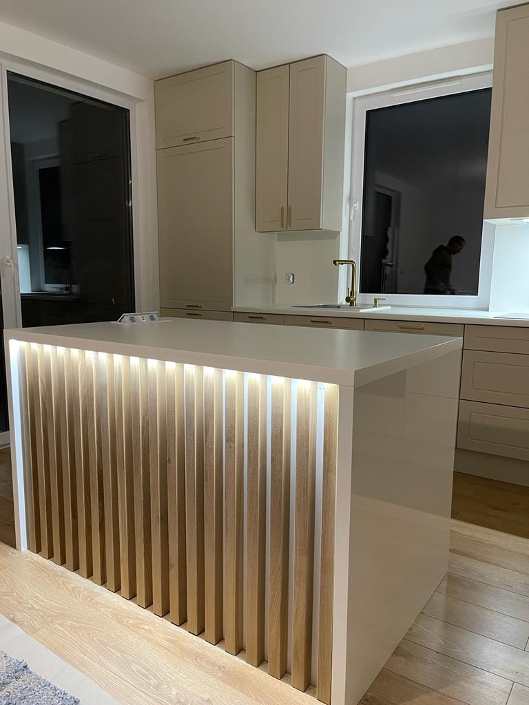 Nowoczesna wyspa kuchenna z oświetleniem LED w pionowych panelach drewnianych, beżowe szafki kuchenne w tle, parkiet na podłodze.