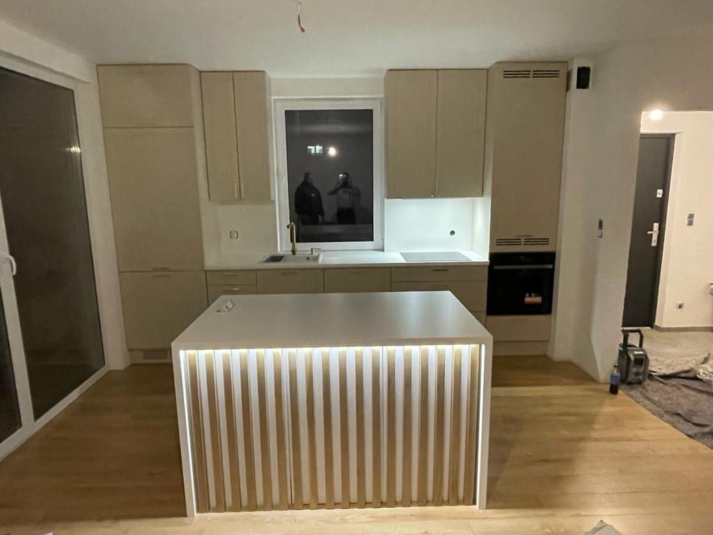 Jasna kuchnia z wyspą oświetloną od dołu, okno z widocznymi sylwetkami osób na zewnątrz, parkiet na podłodze.