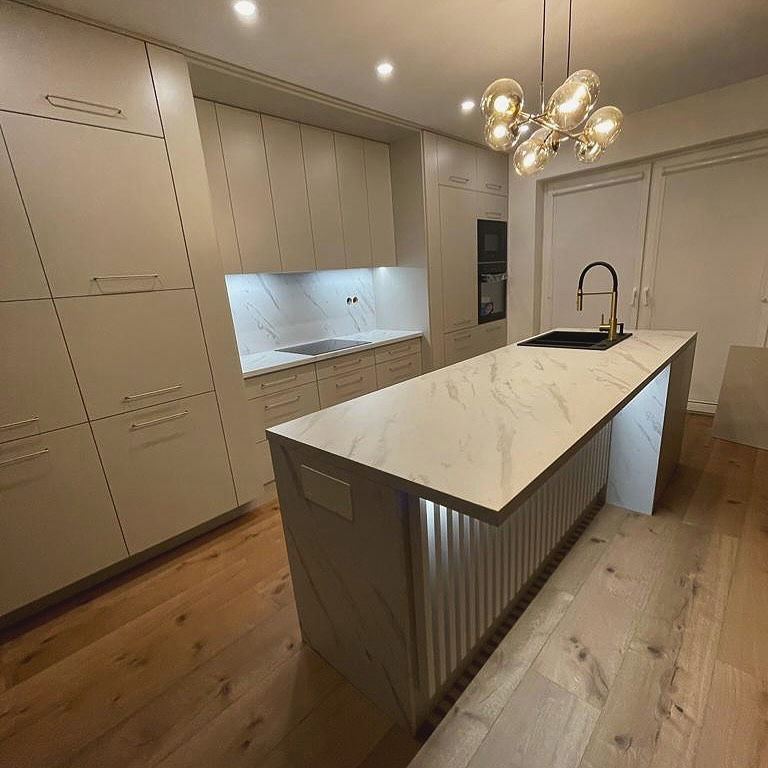 Jasna kuchnia z wyspą, marmurowy blat, złota bateria i designerska lampa wisząca z kulistymi kloszami. Fronty szafek gładkie, bez uchwytów, podświetlenie LED pod wyspą.