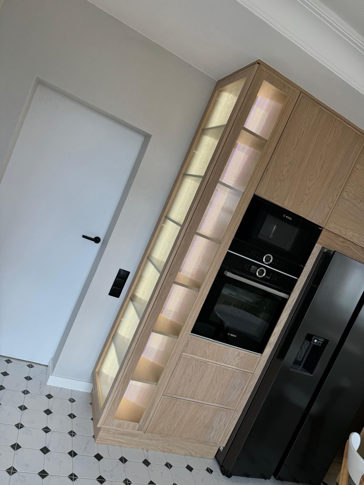 Jasna, dębowa zabudowa kuchenna z podświetlanymi półkami, piekarnikiem i lodówką marki Bosch, na tle białej ściany i podłogi w czarno-białą szachownicę.