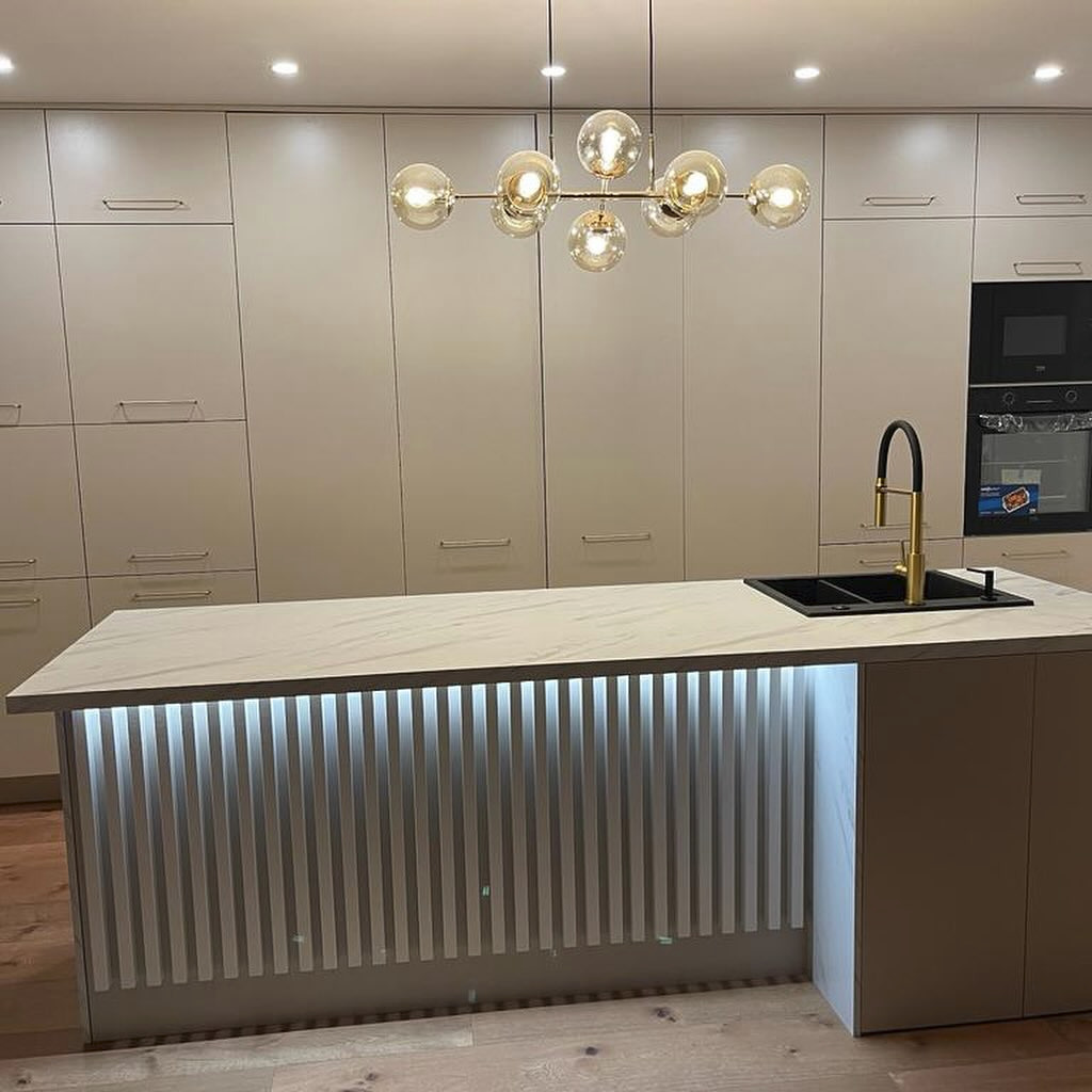Nowoczesna kuchnia z białymi, gładkimi frontami szafek, wyspą z podświetleniem LED i marmurowym blatem, oraz złotą armaturą.