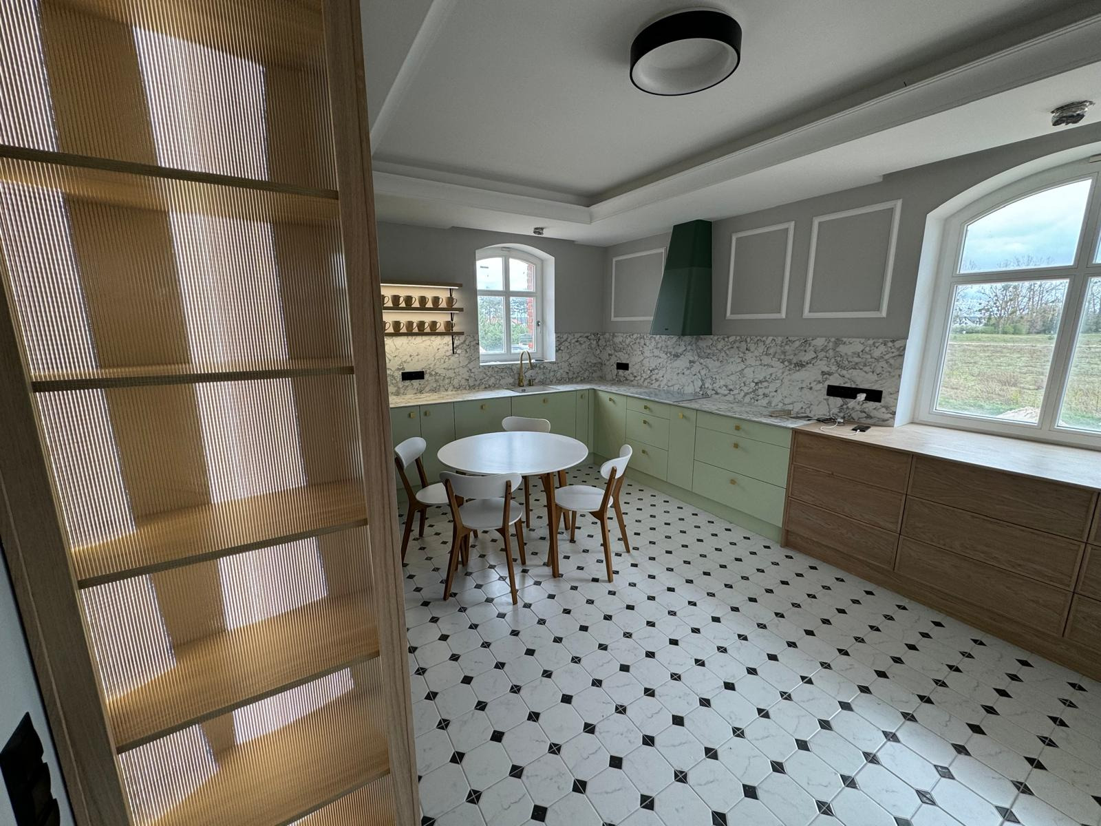 Jasna kuchnia z zielonymi szafkami, marmurowym blatem, białym okrągłym stołem i wzorzystą podłogą, widok z perspektywy otwartego regału.