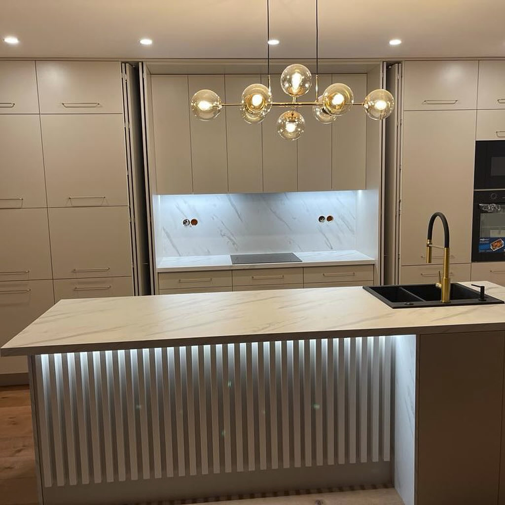 Nowoczesna kuchnia z beżowymi szafkami na wymiar, wyspą z podświetleniem LED i złotą armaturą, oświetlona designerskim żyrandolem z kulistymi kloszami.
