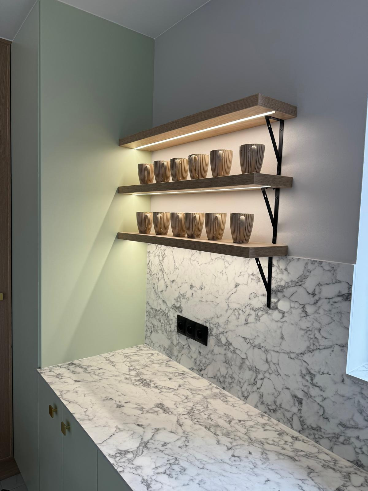 Nowoczesna kuchnia z marmurowym blatem i podświetlanymi półkami, na których stoją identyczne kubki w paski. Widoczne czarne gniazdka na ścianie.