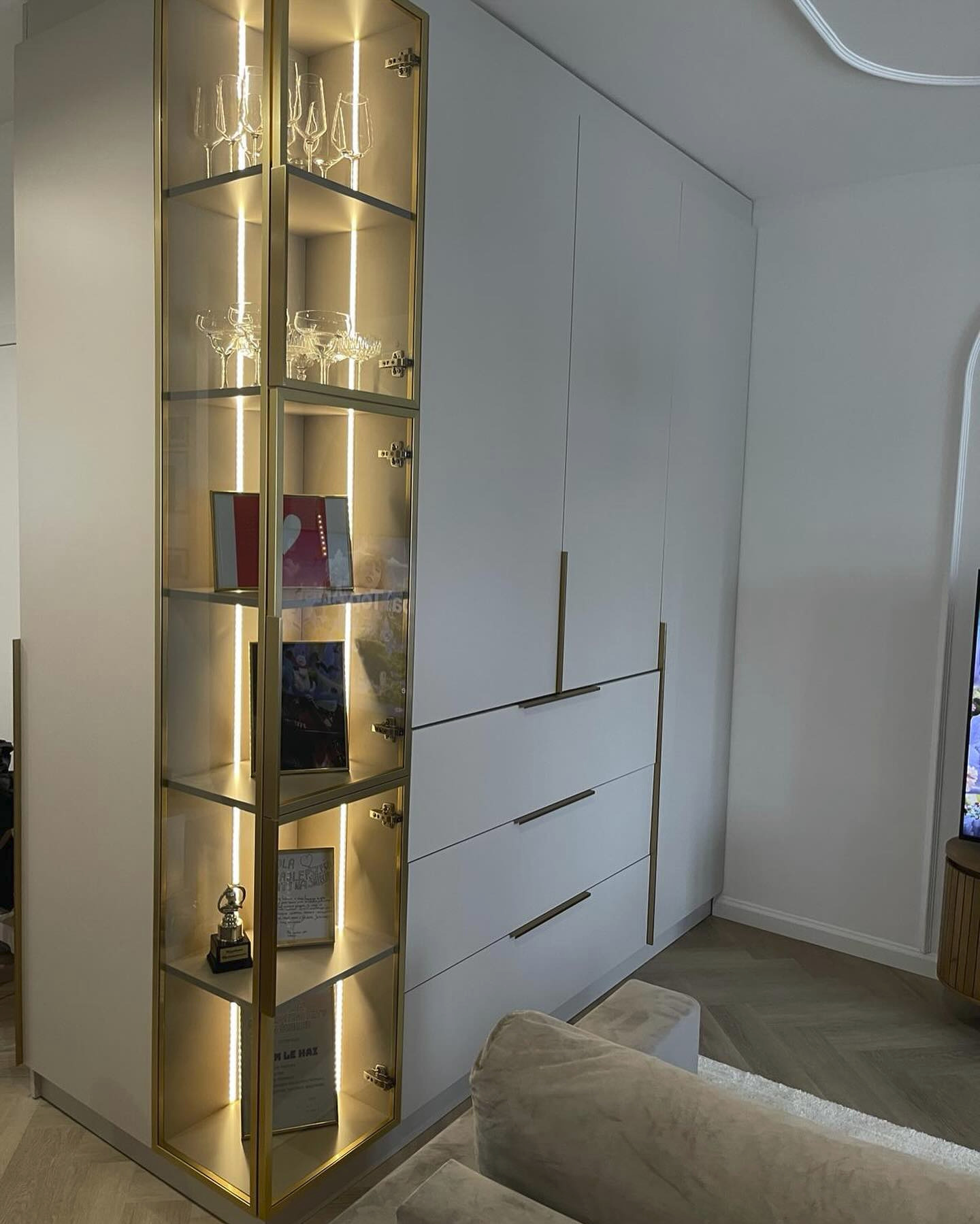 Biała szafa na wymiar z przeszkloną witryną oświetloną ledami, eksponująca szkło, nagrody i zdjęcia, z widocznymi długimi, poziomymi uchwytami.