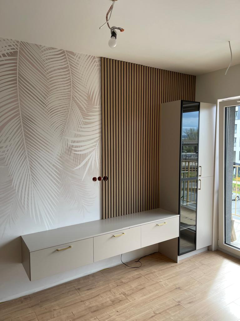 Beżowa szafa wnękowa z lustrzanymi drzwiami, zamontowana pod drewnianą ścianką działową z pionowymi listwami i tapetą w liście palmowe, widok na balkon za oknem.