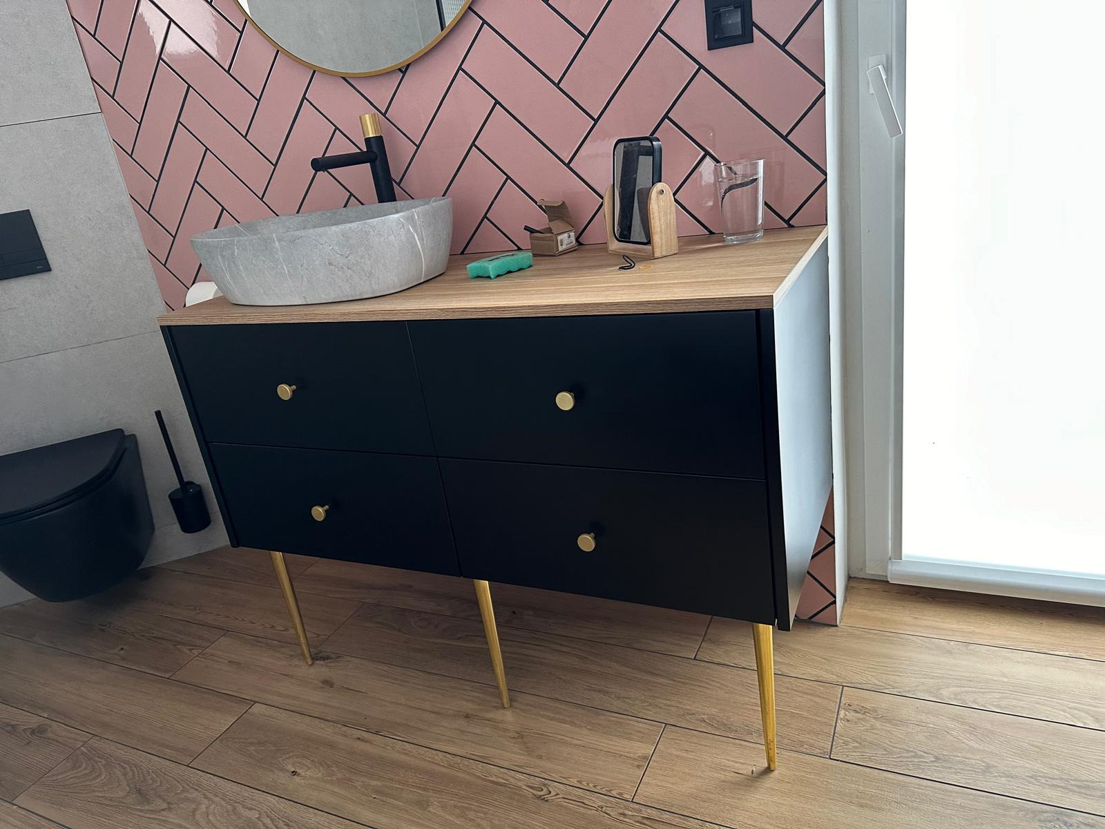 Czarna szafka łazienkowa z czterema szufladami na złotych nóżkach, z umywalką w kształcie kamienia, na tle różowych płytek ułożonych w jodełkę, z widocznym fragmentem toalety i akcesoriów.